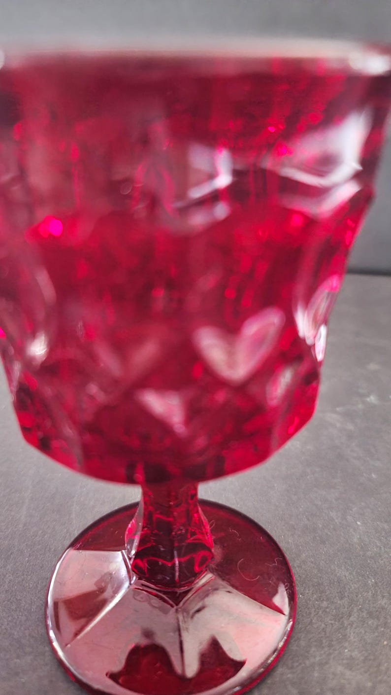 Op de afbeelding: Een robijnrode glazen beker met een hartvormig patroon. De steel en voet zijn ook rood en het glas heeft een textuur. De beker is waarschijnlijk bedoeld voor het serveren van dranken.
