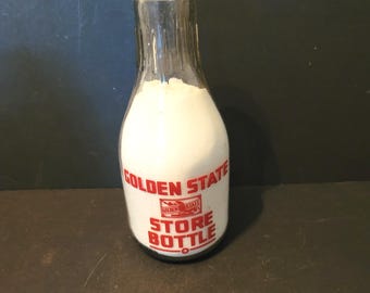 Puede incluir: Una botella de leche de vidrio transparente con interior blanco. La botella tiene un cuello estrecho y una base más ancha. El texto rojo en la botella dice "GOLDEN STATE STORE BOTTLE". La botella está llena de una sustancia blanca, posiblemente leche.