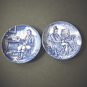 May include: Two blue and white decorative plates. Each plate features a detailed illustration of a seated man in a room setting. One plate is labeled "Huntsman" and the other "Pedlar". The plates have a scalloped edge.