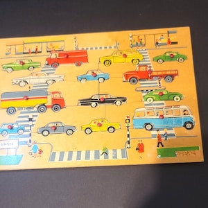 May include: A rectangular wooden peg puzzle featuring a colorful city street scene with various vehicles including cars, a truck, a bus, and a van. Each vehicle has a small red knob for handling. The word SIMPLEX is printed in the bottom left corner.