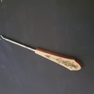 Peut inclure: Un crochet vintage avec un manche décoratif. Le manche est de couleur crème avec une bordure marron et un motif doré orné. Le crochet est en métal.