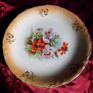 May include: A vintage porcelain plate with a floral design. The plate features a central bouquet of red and pink flowers with green leaves. The rim has a gold-toned trim and decorative floral accents.