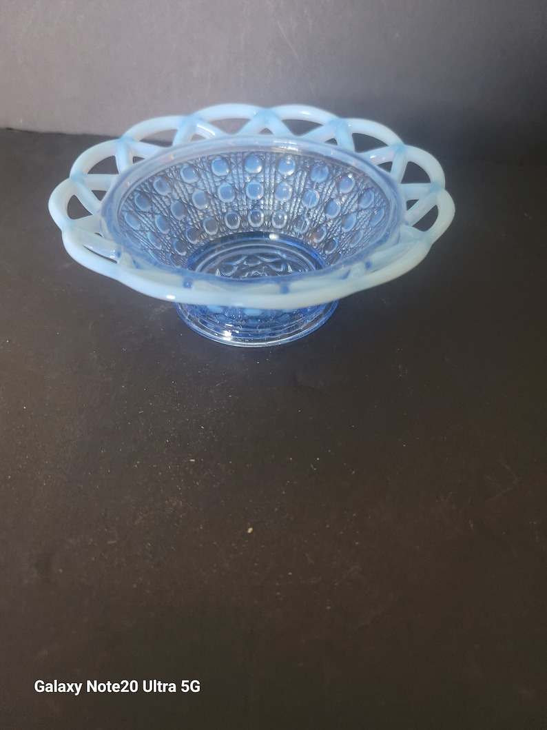 May include: A light blue, vintage glass basket with a delicate, openwork rim. The interior features a textured pattern of raised circles. The basket sits on a small, round base. The overall design is elegant and decorative.