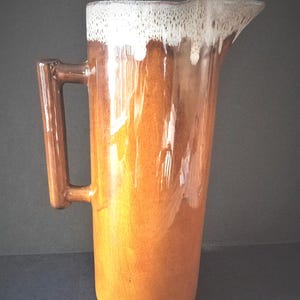 May include: Tall, vintage ceramic pitcher with a brown and tan glaze. The pitcher has a handle and a spout. The top edge has a white, dripped glaze. A classic piece for serving beverages.