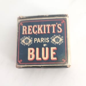 May include: A vintage box of Reckitt's Paris Blue. The box is rectangular with a dark blue background and red trim. The text "RECKITT'S PARIS BLUE" is displayed in a cream-colored font. The box has a regal design with a crown.