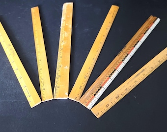 Puede incluir: Una colección de reglas de madera vintage de varias longitudes, dispuestas sobre una superficie oscura. Las reglas son principalmente de madera amarillenta, con marcas negras y rojas que indican centímetros. Algunas muestran signos de desgaste.