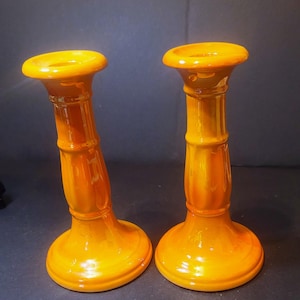 Pode incluir: Dois castiçais de cerâmica laranja. Cada um tem uma base redonda, um caule cilíndrico com ranhuras verticais e um topo circular. Os castiçais são de cor laranja brilhante e estão sobre um fundo escuro.