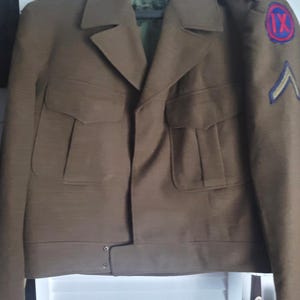Puede incluir: Chaqueta militar marrón con dos bolsillos delanteros y cuello con solapas. La chaqueta tiene un parche en la manga con el número IX en un círculo rojo y un galón azul. La chaqueta está colgada en una percha.