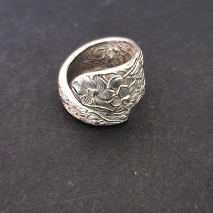Puede incluir: Un anillo plateado con un diseño floral. El anillo parece estar hecho de una cuchara, con el patrón floral envolviendo la banda. El anillo se encuentra sobre un fondo oscuro.