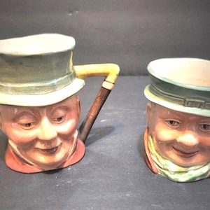 Puede incluir: Dos tazas de cerámica con forma de caras sonrientes que llevan sombreros. Una taza tiene un sombrero de copa con un asa de bastón, y la otra una gorra. Las tazas están pintadas con tonos de piel, acentos rojos, verdes y amarillos.