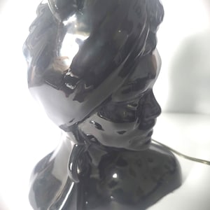 May include: A black ceramic bust of a person with a head covering. The bust has a glossy finish and a detailed facial profile. The figure's neck and shoulders are also visible, and a power cord extends from the base.