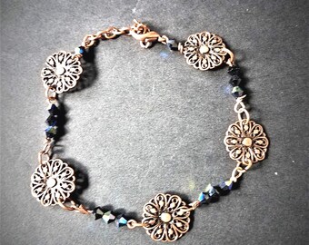 Könnte beinhalten: Ein kupferfarbenes Armband mit filigranen, blütenförmigen Anhängern und dunkelblauen Kristallperlen. Das Armband ist durch eine filigrane Kette verbunden und hat einen Verschluss. Der Schmuck wird auf einer dunklen Oberfläche präsentiert.