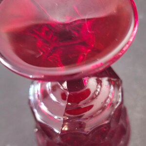 Op de afbeelding: Een robijnrode glazen kelk met een facetgeslepen steel en een ronde kom. Het glas heeft een glanzende afwerking en staat tegen een donkere achtergrond. De kelk is waarschijnlijk bedoeld voor het serveren van wijn of andere dranken.