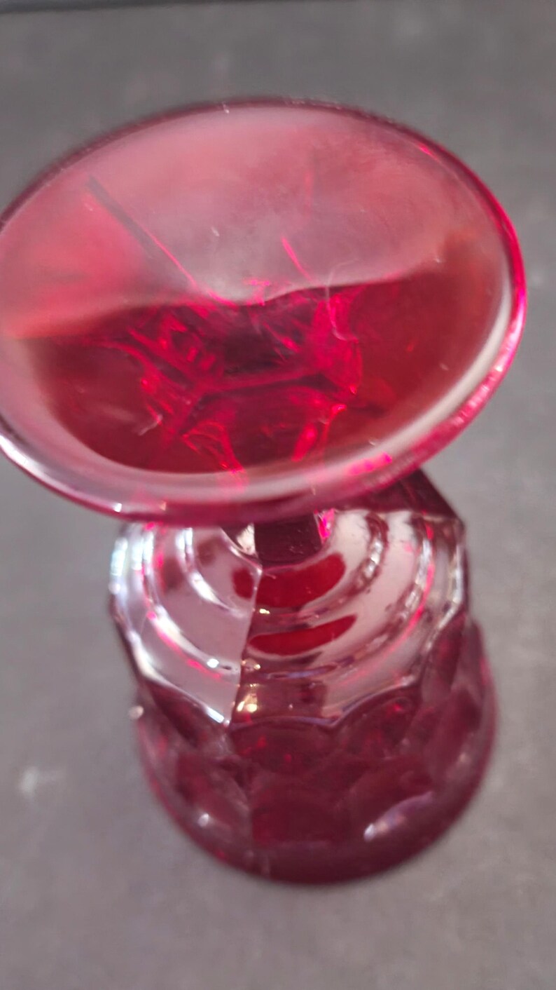 Op de afbeelding: Een robijnrode glazen kelk met een facetgeslepen steel en een ronde kom. Het glas heeft een glanzende afwerking en staat tegen een donkere achtergrond. De kelk is waarschijnlijk bedoeld voor het serveren van wijn of andere dranken.
