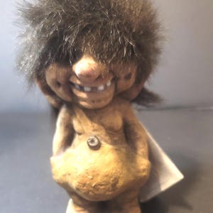 Puede incluir: Una figura de muñeca troll vintage con una cara sonriente, dientes visibles y cabello castaño y difuso. La muñeca viste un atuendo color canela con un solo botón. Una etiqueta blanca está adherida a la espalda.