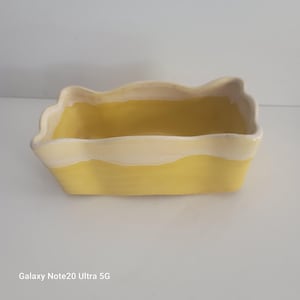 May include: A rectangular ceramic planter with a wavy rim. The planter is painted a bright yellow with a cream-coloured rim. The planter is empty and sits on a white surface.
