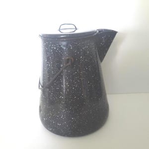 Puede incluir: Una cafetera vintage de esmalte negro y blanco moteado, con tapa y asa. La cafetera tiene un pico para verter y un asa de metal. El patrón moteado le da una estética rústica y de granja.
