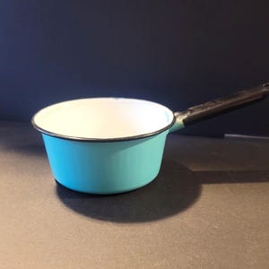Peut inclure: Petite casserole turquoise avec une poignée noire. L'intérieur est blanc, et le bord est souligné de noir. La casserole est posée sur une surface sombre, projetant une ombre. Article de cuisine vintage.