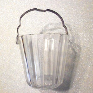 May include: A clear, ribbed glass bucket with a dark metal handle. The bucket has a flared shape and vertical lines. The handle is arched and attached to the bucket's rim. The bucket is likely for decorative purposes.