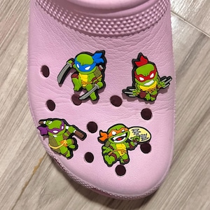 May include: A light pink clog shoe adorned with four colorful Teenage Mutant Ninja Turtles shoe charms. The charms feature the turtles in action poses, each with their signature weapons and masks. The shoe has multiple ventilation holes.