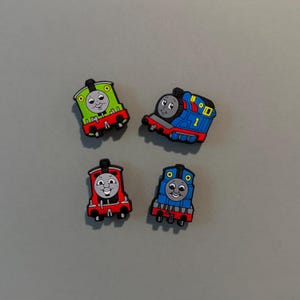 May include: Four colorful train-shaped charms. The charms feature smiling faces and are in green, blue, red, and blue. Each train has a unique design, with details like wheels and smokestacks. These charms are likely for crafts or accessories.