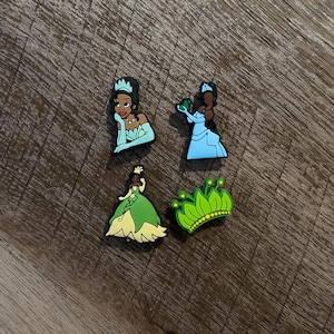 May include: Four colorful shoe charms featuring princess designs. The charms include a princess in a light blue dress, a princess in a green dress, a princess with a crown, and a green crown charm.