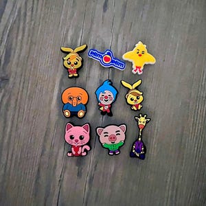 May include: A set of ten colorful cartoon character shoe charms. The charms feature a variety of characters, including a yellow chick, a pink pig, a blue clown, a purple giraffe, a pink cat, a yellow bunny, a brown elephant, and a yellow character with a red nose. The charms are all about 2 inches tall and are made of a soft, flexible material.
