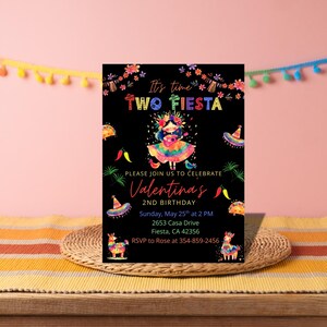 Op de afbeelding: Een zwarte uitnodigingskaart met de tekst "It's time TWO FIESTA" en "PLEASE JOIN US TO CELEBRATE Valentina's 2ND BIRTHDAY". De kaart bevat kleurrijke illustraties van een meisje, sombrero's en andere feestelijke elementen. De kaart ligt op een geweven mat.