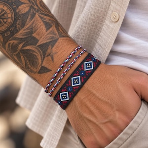 May include: A set of three bracelets consisting of two thin braided cords in red, white, and blue, and one wider black woven band featuring a repeating geometric pattern in red, white, and blue diamond shapes with small central blue circles.
