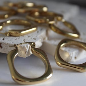 May include: Close-up of several gold-colored hoop earrings. The earrings have a square shape with rounded edges. The earrings are on a white surface and a textured, off-white surface. The earrings are a shiny, metallic gold color.