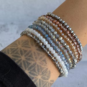 May include: A collection of beaded bracelets in various colors, including white, gray, blue, and brown. The bracelets are stacked on a wrist, showcasing the different shades and textures of the beads. The bracelets are a fashion accessory.