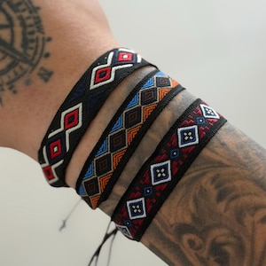 May include: Three woven fabric bracelets featuring intricate geometric patterns. The first bracelet displays red and white diamond shapes on a black background. The second features orange, blue, and brown diamonds. The third has red, white, and blue accents.