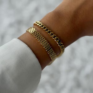 Puede incluir: Dos pulseras doradas adornan una muñeca, una con un diseño de cadena de eslabones y la otra con un patrón de malla. Las pulseras, en un tono dorado brillante, se llevan sobre una camisa blanca de manga larga.