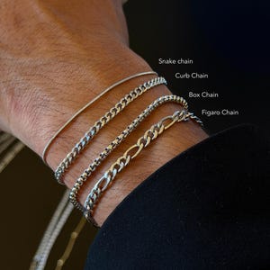 Może przedstawiać: Cztery srebrne bransoletki łańcuszkowe: łańcuszek wężyk, łańcuszek typu curb, łańcuszek typu box i łańcuszek Figaro. Bransoletki są prezentowane na nadgarstku, prezentując różne wzory i tekstury każdego łańcuszka. Obraz jest dobrze oświetlony.