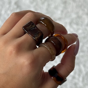 Könnte beinhalten: Eine Hand, die eine Sammlung von Harzringen in verschiedenen Braun- und Bernsteintönen zeigt. Die Ringe haben unterschiedliche Formen, darunter ein rechteckiger Ring mit Marmormuster und ein runder Ring mit Schildpattmuster.
