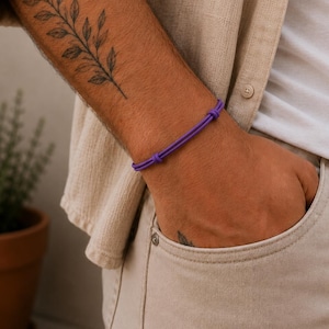 May include: A simple purple cord bracelet featuring two sliding knots for adjustable sizing. The bracelet consists of a single smooth, vibrant purple string that wraps around the wrist, secured by two small knots that allow the diameter to be changed.