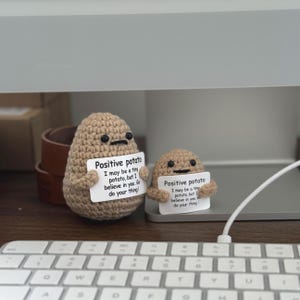 Puede incluir: Dos figuras de patata de ganchillo, una grande y otra pequeña, cada una con un cartel que dice "Positive potato". Los carteles incluyen el texto "I may be a tiny potato, but I believe in you. Go do your thing!" Las patatas son de color beige.