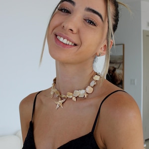 May include: A seashell necklace with a gold starfish pendant. The necklace is made of various sizes and shapes of off-white seashells. The model is wearing a black spaghetti strap top.