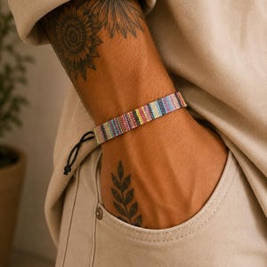 May include: A woven fabric bracelet featuring a multi-colored striped pattern with thin vertical lines in shades of pink, blue, yellow, and white. The bracelet includes a black cord closure with a small knot at the end for an adjustable fit.