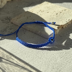 May include: A blue woven bracelet with an evil eye charm. The bracelet is made of a woven cord and features a small, round charm with a blue eye design. The bracelet is adjustable and has a silver-colored bead at the end.