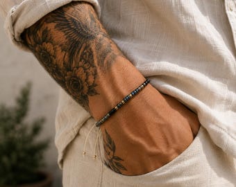 May include: A thin bracelet featuring small, alternating dark metallic and gold-toned beads arranged in a repeating pattern. The bracelet includes a light-colored cord with two dangling ends, each finished with a tiny gold-toned bead at the tip.