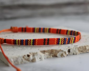 Puede incluir: Pulsera de la amistad tejida naranja con un patrón de rayas en azul, amarillo, rojo y negro. La pulsera está hecha a mano y tiene un cierre ajustable. La pulsera es un accesorio de estilo bohemio.