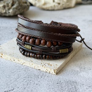 Puede incluir: Una pulsera de cuero marrón multicapa con cuentas de madera y detalles metálicos. La pulsera presenta varias texturas, incluyendo cuero trenzado y cuentas de madera lisas. El diseño es casual y rústico.
