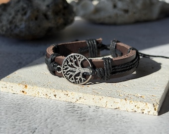May include: A brown leather bracelet with a silver tree of life charm. The bracelet features black braided accents and a clasp closure. The jewelry is displayed on a light-colored, textured surface.