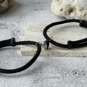 May include: Two black rope bracelets with a heart-shaped charm. The bracelets have a magnetic clasp and are displayed on a light-coloured stone surface. The heart charm is silver and black.