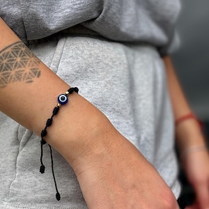 May include: Black string bracelet featuring a blue and white evil eye charm with gold-colored beads. The bracelet is knotted and adjustable, designed to be worn on the wrist. The bracelet is shown on a person's arm.