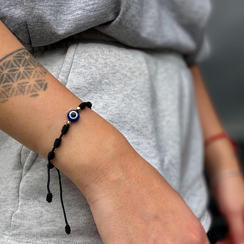 May include: Black string bracelet featuring a blue and white evil eye charm with gold-colored beads. The bracelet is knotted and adjustable, designed to be worn on the wrist. The bracelet is shown on a person's arm.