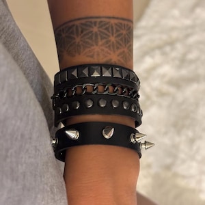 Puede incluir: Un conjunto de cuatro pulseras de cuero negro con tachuelas y púas plateadas. Las pulseras están apiladas en la muñeca de una persona.