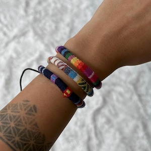 May include: Three colorful woven bracelets stacked on a person's wrist. The bracelets are made of different colored threads and have a variety of patterns.