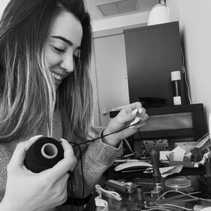 May include: A black spool of thread held in hands with a thin black string extending toward a metal wax seal stamp. The scene includes a white measuring tape, a lighter, and various crafting supplies on a desk in a black and white photograph.